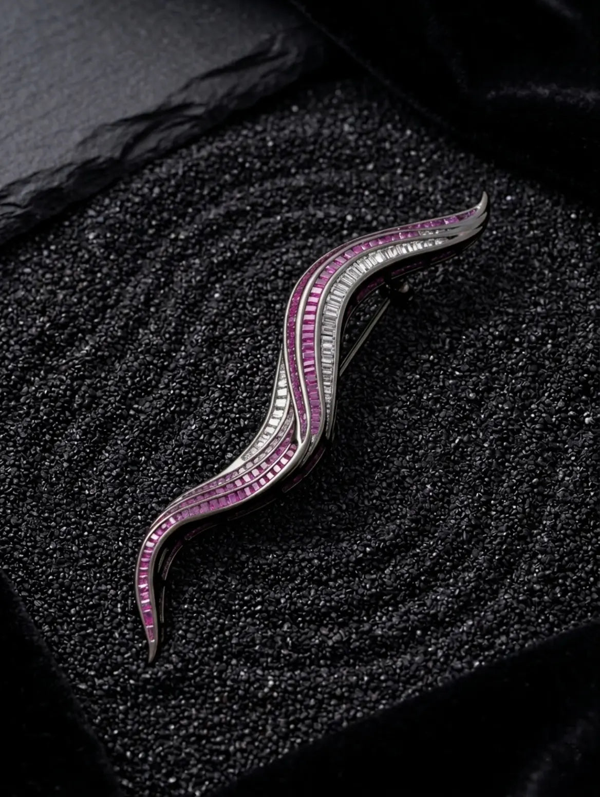 A curved silver-toned brooch or hair pin with rows of pink gemstones and clear stones, displayed on a textured black surface.