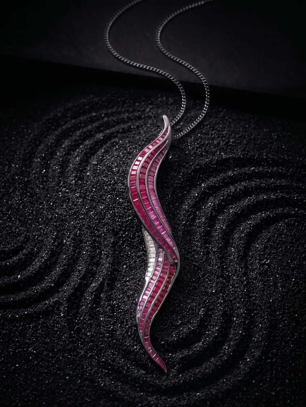 A silver wavy pendant necklace adorned with rows of pink and white gemstones, displayed on a textured black background.