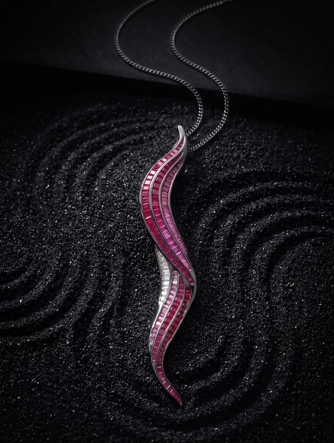 A silver wavy pendant necklace adorned with rows of pink and white gemstones, displayed on a textured black background.