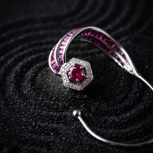 A silver-toned open cuff bracelet featuring a central hexagonal cluster of sparkling clear stones and a prominent round red gemstone, accented by curved rows of deep pinkish-red stones along the inner band, set against a dark textured background.